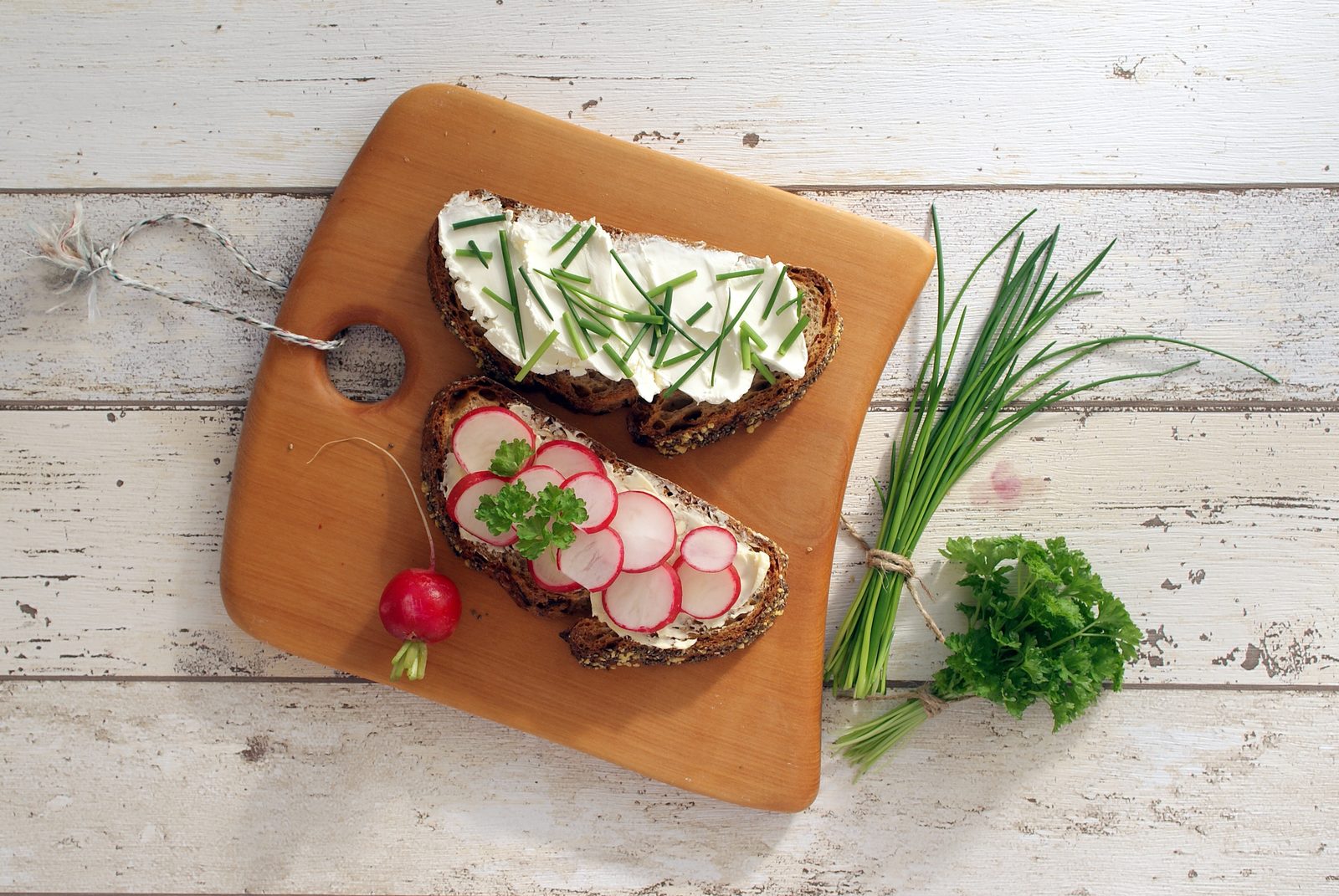 Two slices of dark rye bread topped with biezpiens cottage cheese, fresh chives and sliced radishes on a wooden board