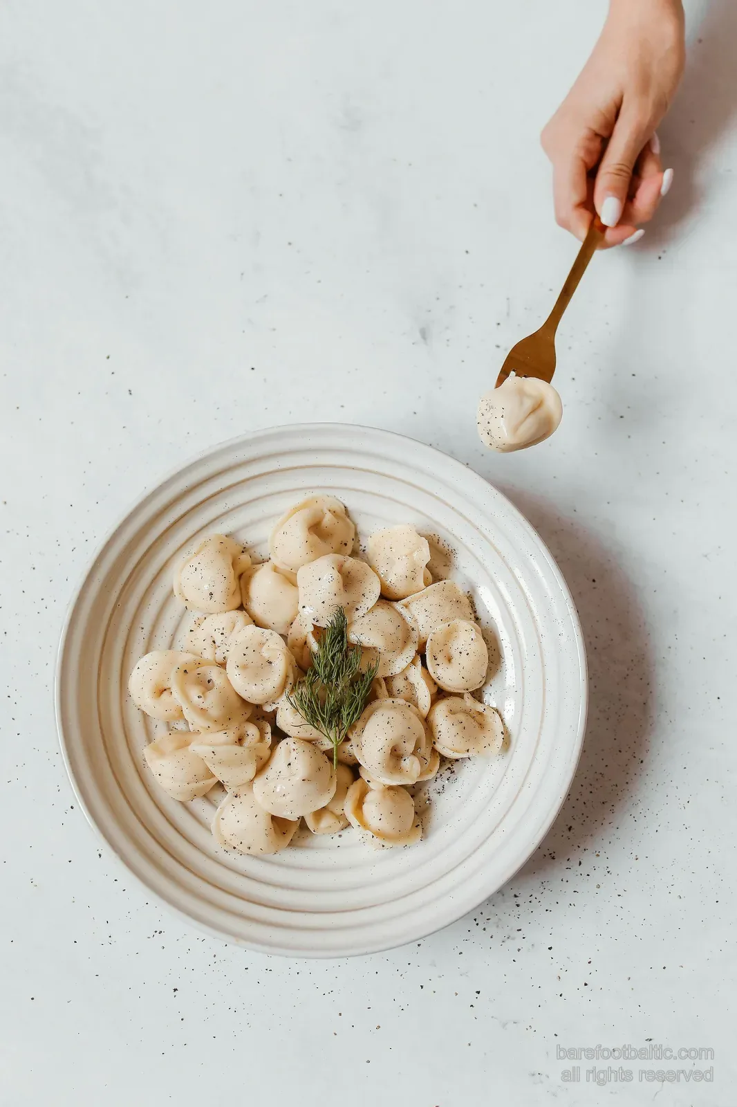 Pelmeņi dumplings on a white plate with a sprig of dill, a hand lifting one with a fork