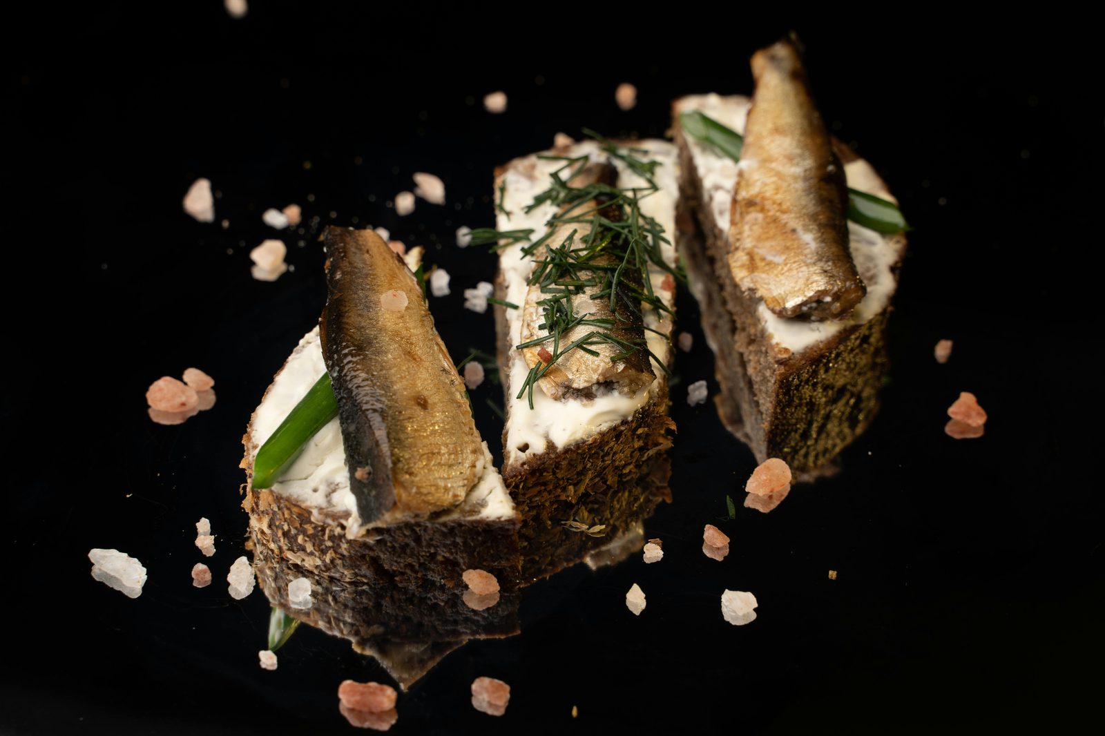 Three smoked sprats on rye bread with sour cream and dill, on a black background